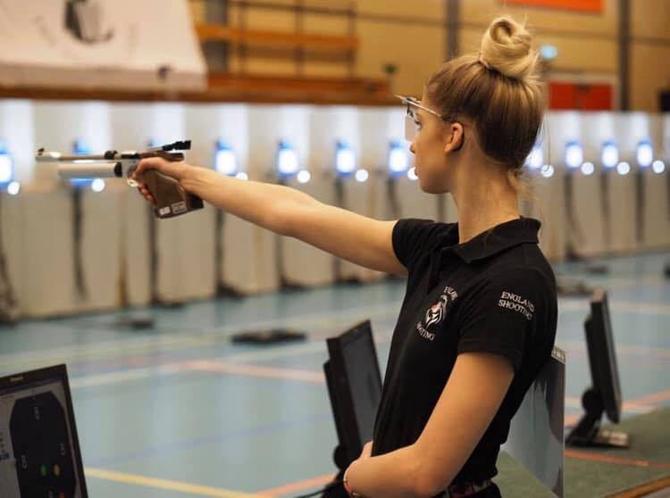 A young lady aims a ten metre match air pistol in competition. She wears a black polo shirt with the words "England Shooting" on the sleeve.