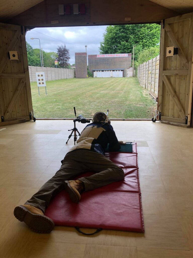 A shooter lies in the prone position in front of an open door looking out onto a rifle range. They shoot a smallbore target rifle through the door onto the outdoor fifty metre range.