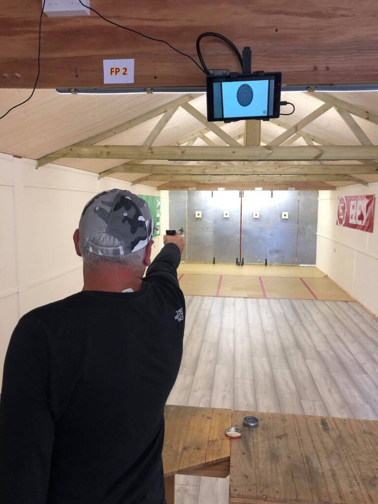 An adult fires a match air pistol at a paper target. Above him, a tablet displays an image of the target from a webcam.