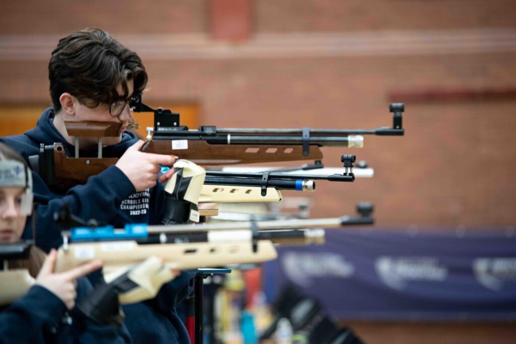 Young competitors shoot match air rifles in a sports hall.