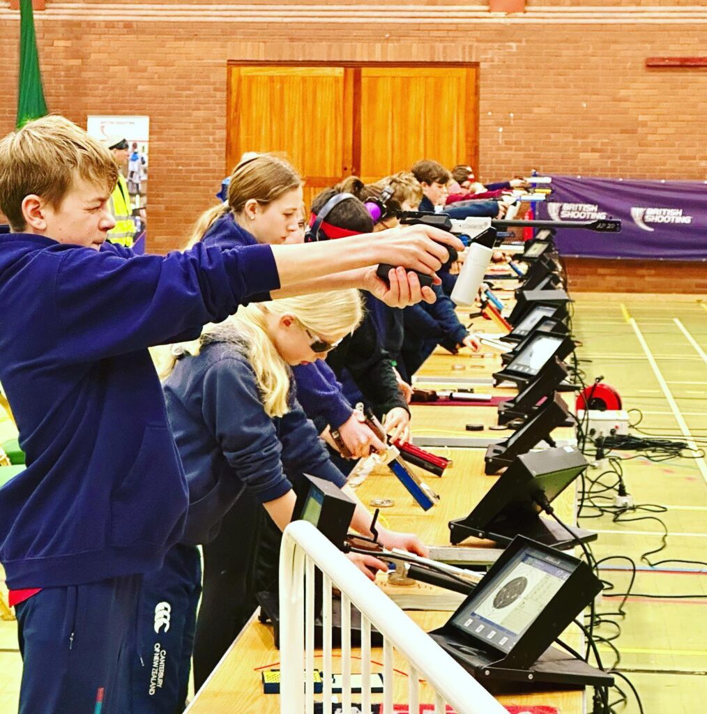 A group of young people fire match air pistols in a sports hall. At the bench in front of them, computer monitors show the scores returned from the electronic targets.