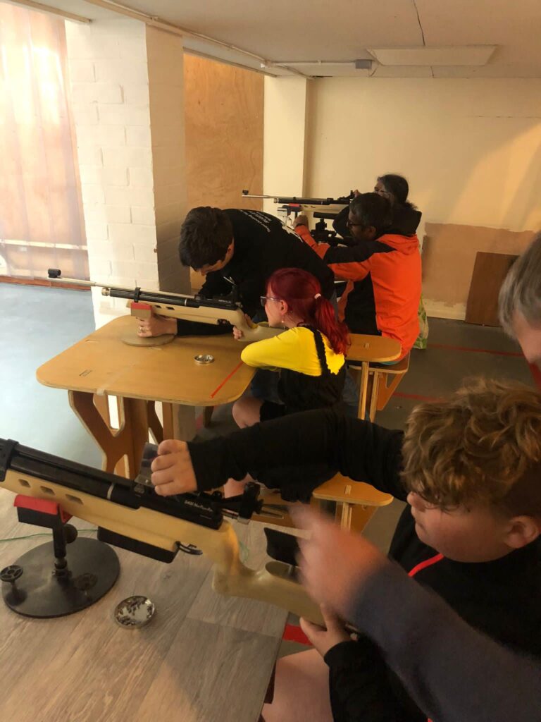 Two young people are sat at benches shooting air rifles from rests.