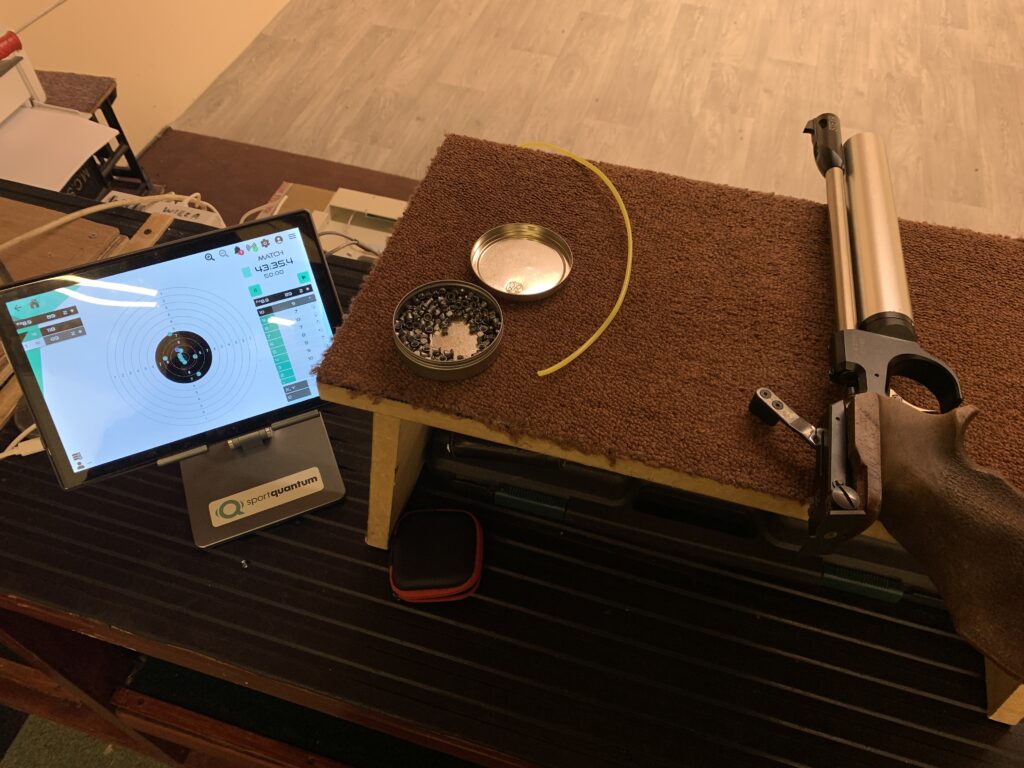 A match air pistol lies on a bench next to a tin of pellets and a tablet computer which displays the shots and scores from an electronic target