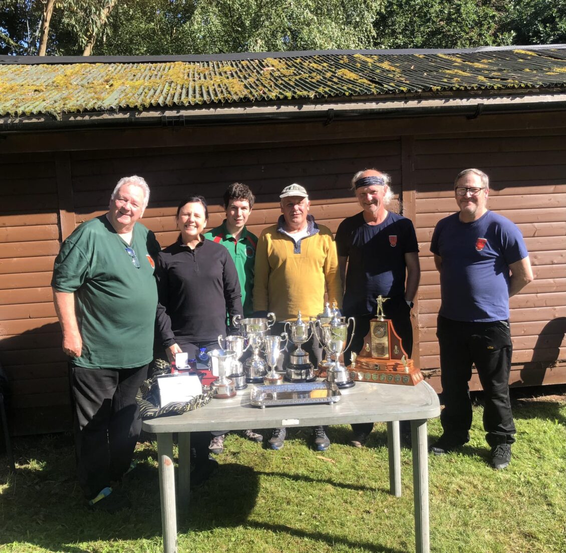 Six competitors stand behind a table of trophies they have won.