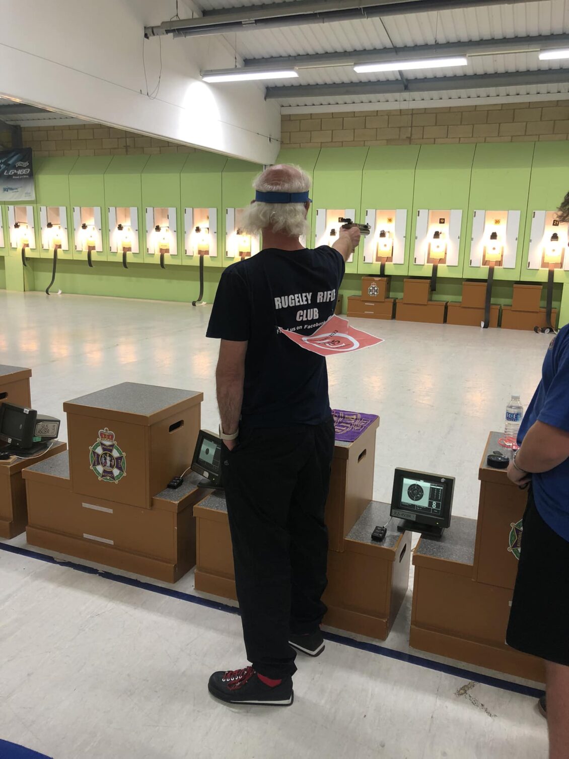 A man fires an air pistol in competition. His back is to the camera, his t-shirt reads "Rugeley Rifle Club"