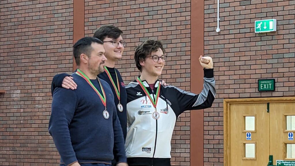 Three medal winning shooters stand on a podium with their medals