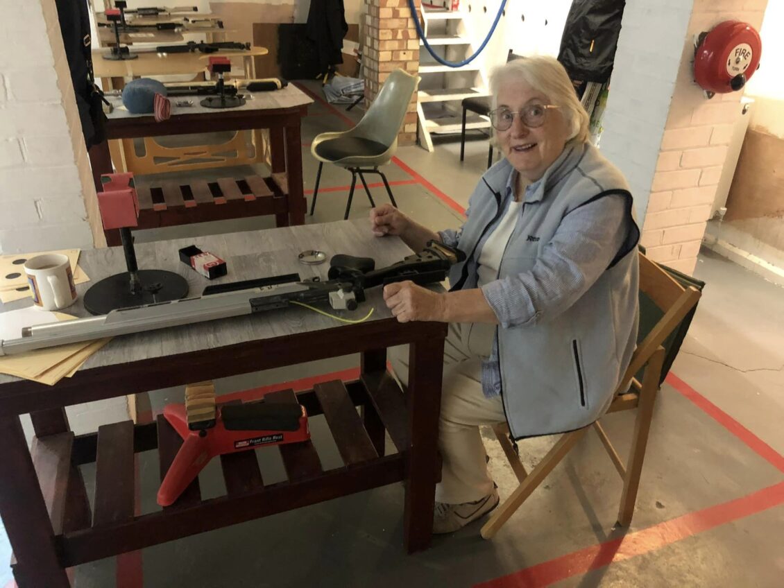 A lady with grey hair and a grey jacket sits at a benchrest table ready to shoot a target air rifle on an indoor shooting range.