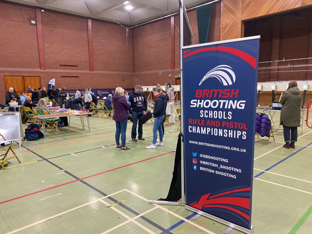 A sports hall built of red brick is configured for target shooting. Electronic targets for air rifles and air pistols are visible in the background. In the foreground, a blue banner identifies the event as the British Shooting Schools Rifle and Pistol Championships.