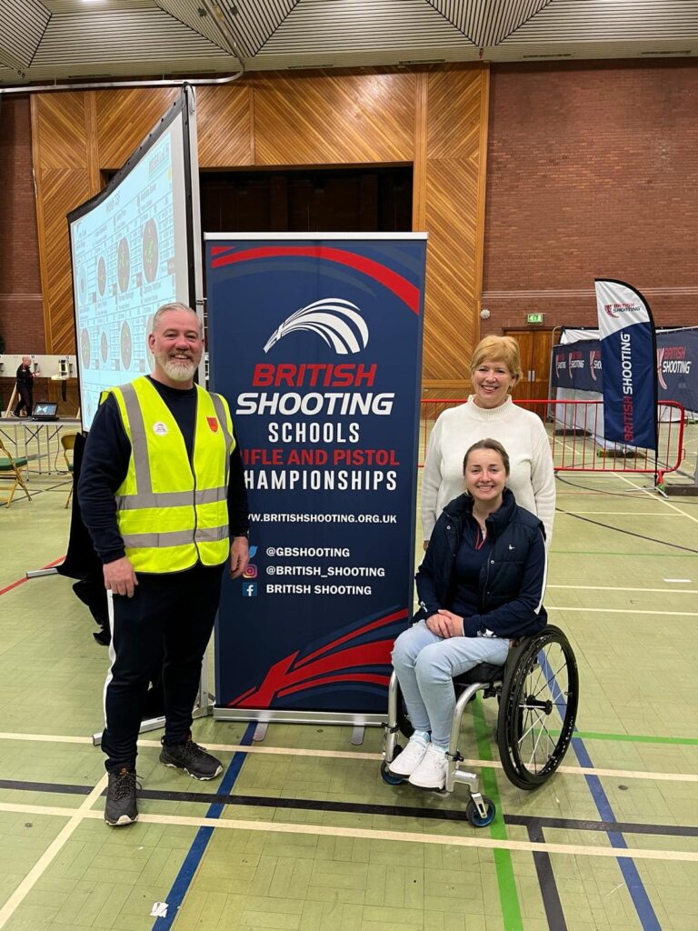 A man in a high-viz jacket is stood with two ladies. One is standing, the other is in a wheelchair. She is Izzy Bailey, Paralympian. The are posed next to a sign identifying the event as the British Shooting Schools Rifle and Pistol Championships