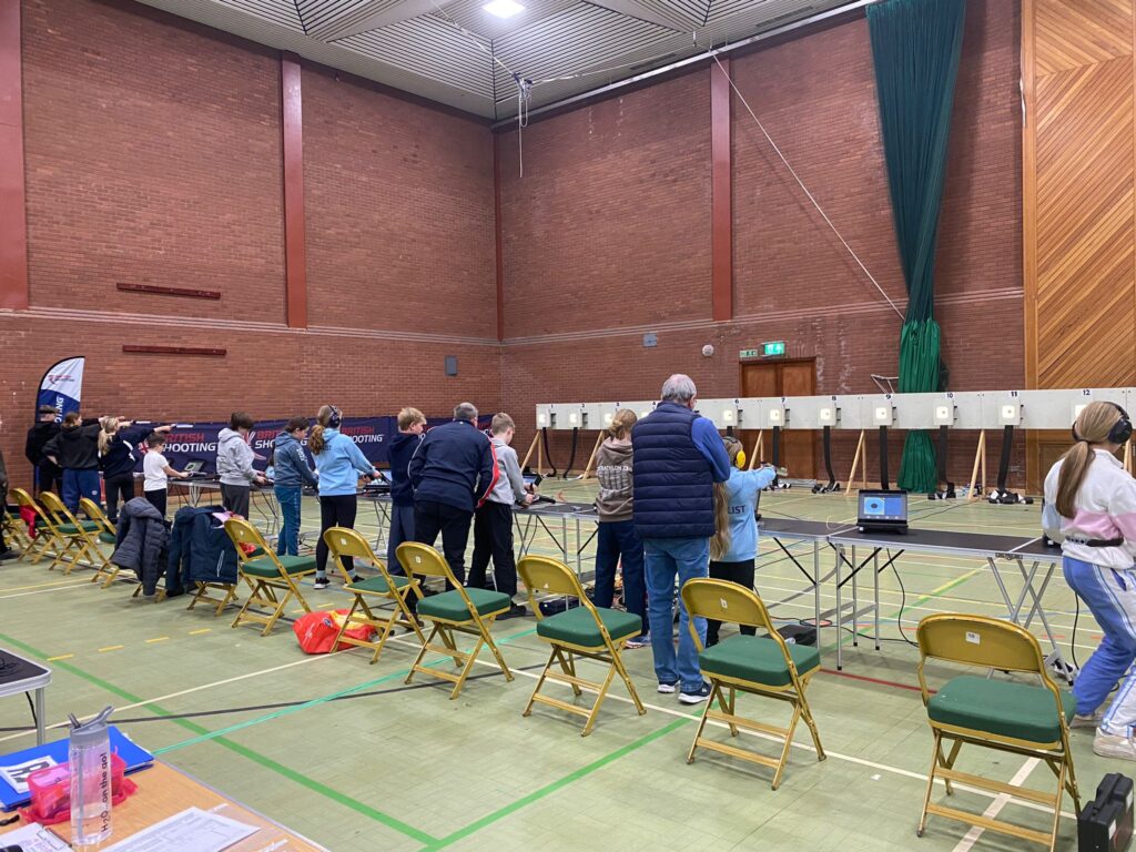 Young people shoot air pistols during a practice session at the British Shooting Schools Rifle & Pistol Championships. The range is located in a brick sports hall set up for the event. Electronic targets are visible in the background with monitors on tables at the firing point.