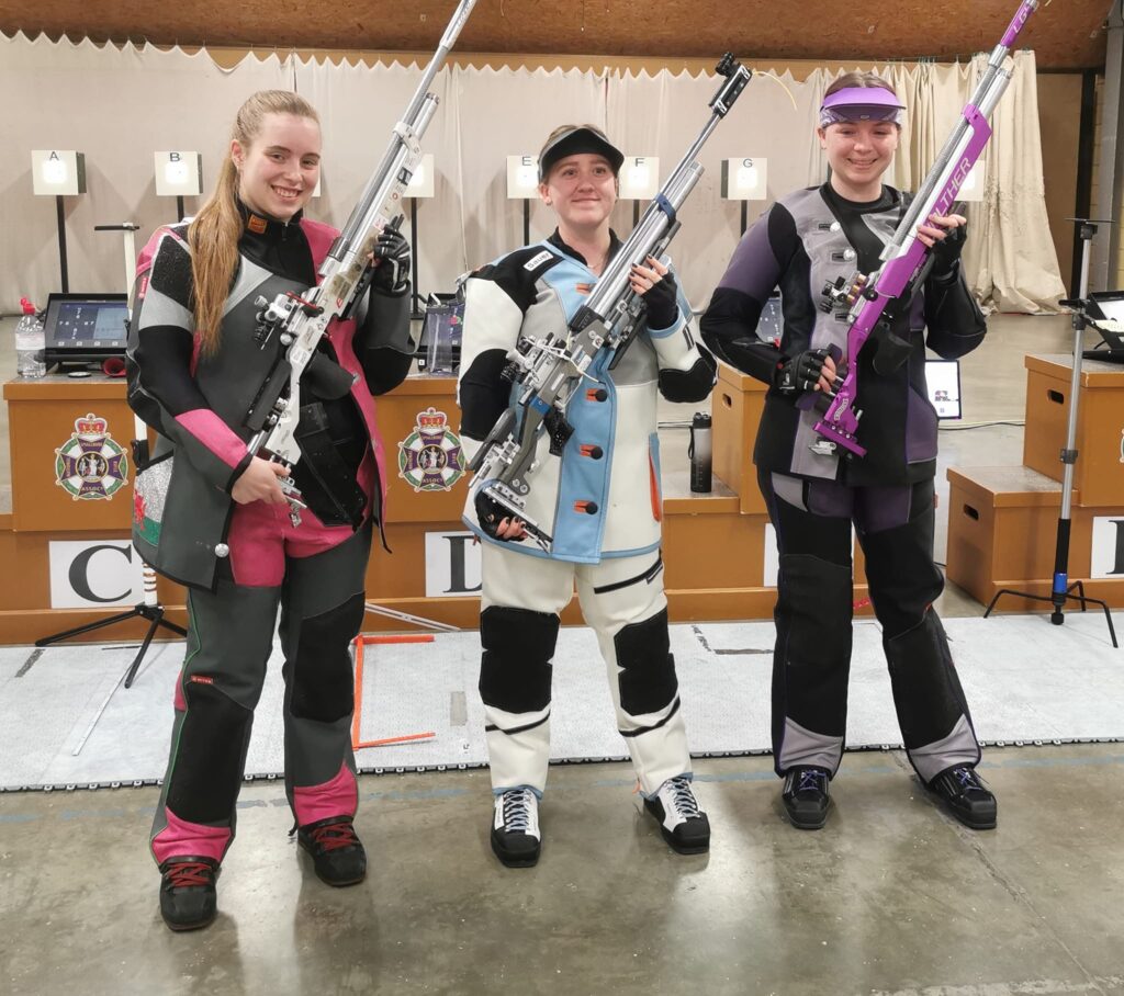 Three young women stand smiling with target air rifles on a finals range.