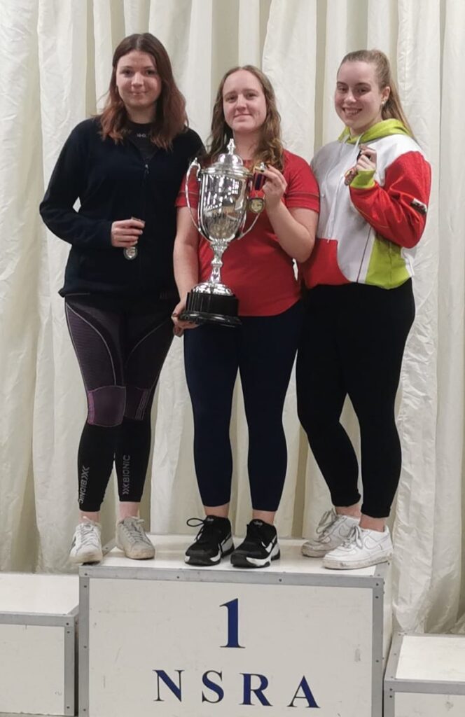 Three smiling women stand on a podium. The athlete in the centre holds a trophy.