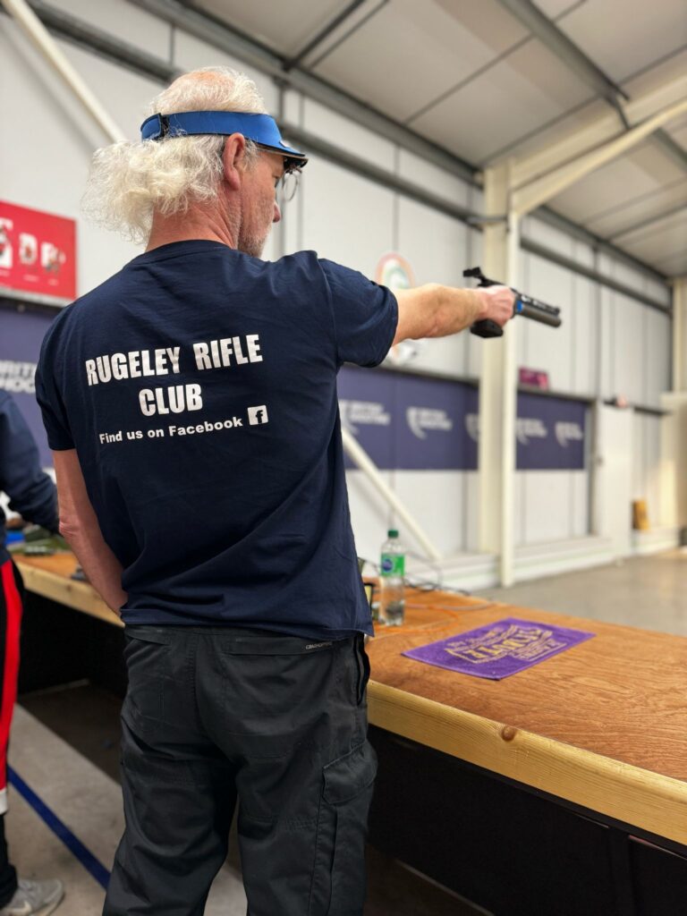 A man aims a target air pistol with his right arm. He is wearing a blue t-shirt with "Rugeley Rifle Club" printed on the back.