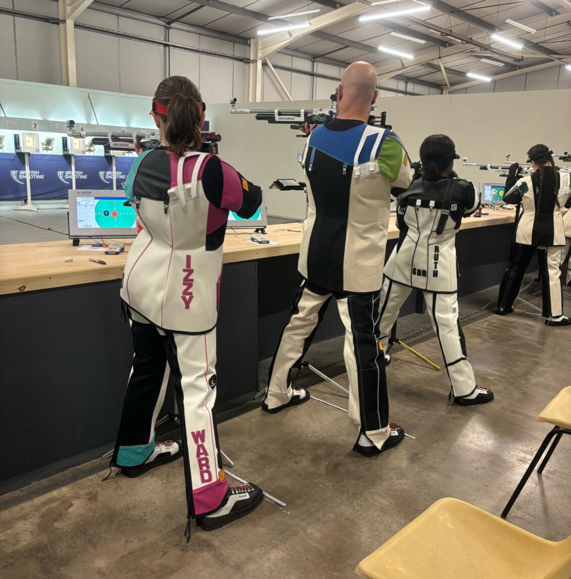 Competitors shoot target air rifles at the British Shooting Air Series competition. Their backs are to the camera and they are wearing special canvas jackets and trousers.