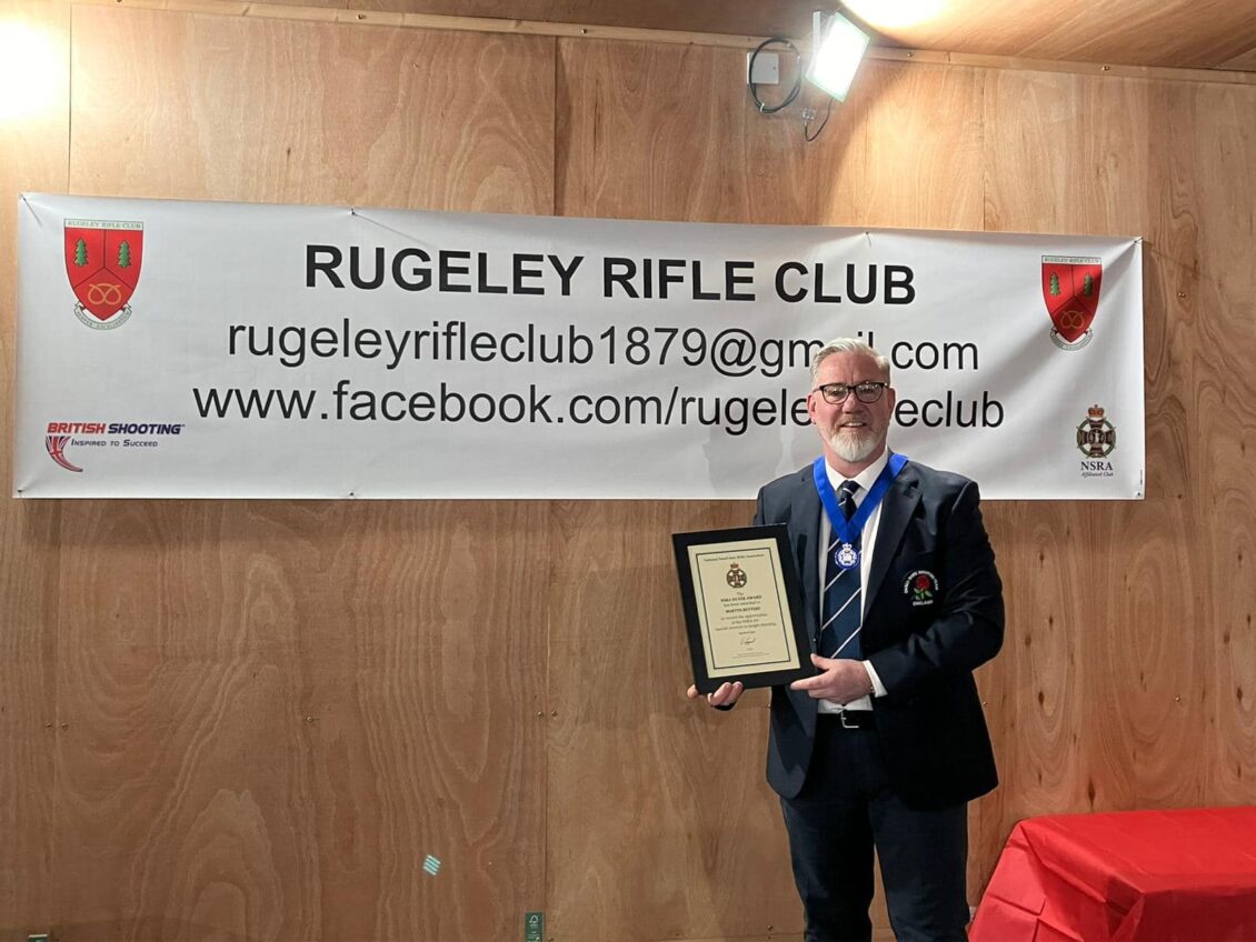 A man stands in front of a banner reading "Rugeley Rifle Club". He is wearing a silver medal on a blue ribbon, and holds a framed certificate recognising his services to target shooting with a Silver Award from the National Smallbore Rifle Association.