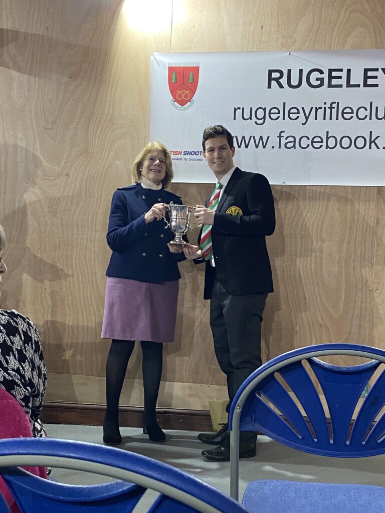 A woman stands on the left at a prize-giving ceremony, presenting a trophy to a man stood on the right.