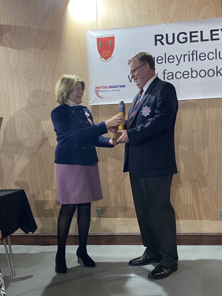 A woman stands on the left at a prize-giving ceremony, presenting a trophy to a man stood on the right.