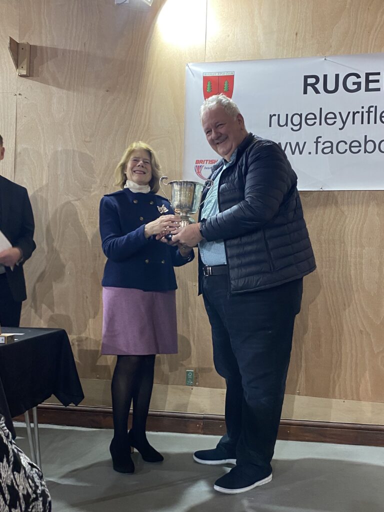 A woman stands on the left at a prize-giving ceremony, presenting a trophy to a man stood on the right.