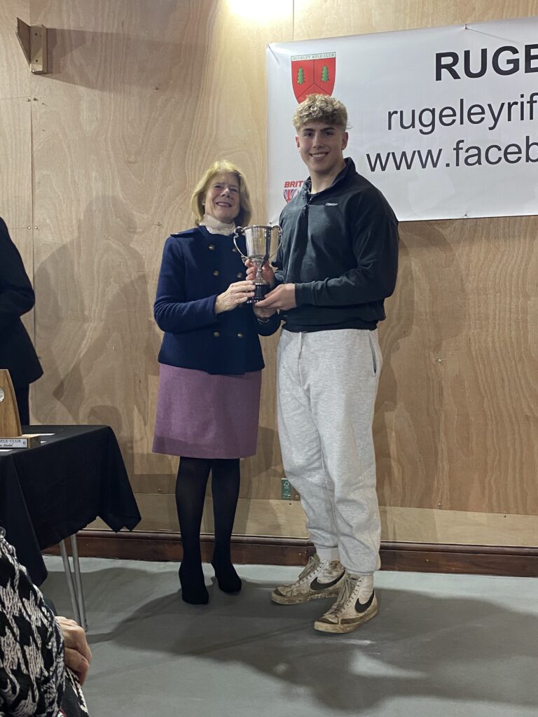 A woman stands on the left at a prize-giving ceremony, presenting a trophy to a young man stood on the right.