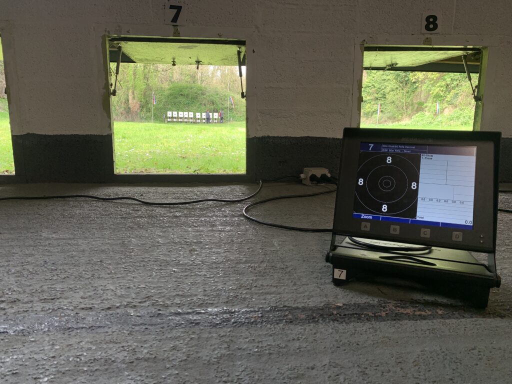 A monitor shows a blank target from an electronic scoring system. Through a hole in the wall, the bank of targets are visible fifty metres away on the outdoor range. Two range officers are just visible inspecting the targets before the next detail starts.