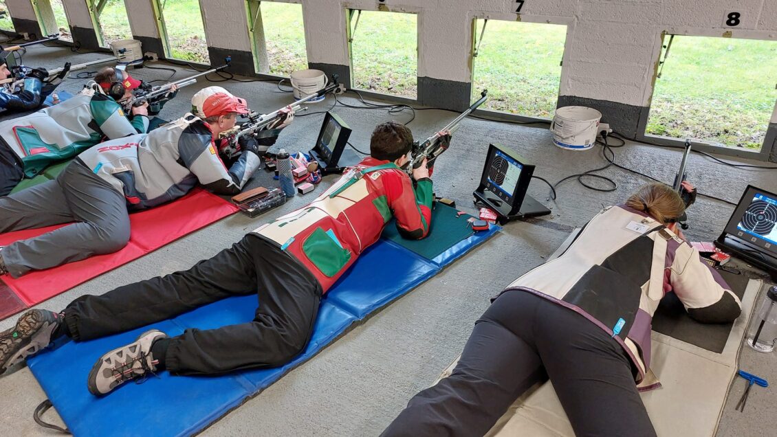 A group of floor shooters lie prone on the floor of an indoor firing point, aiming out through square holes in the wall at targets on the outdoor range. They wear canvas and leather jackets for support, some in bright colours. Monitors show the score from the electronic target system.