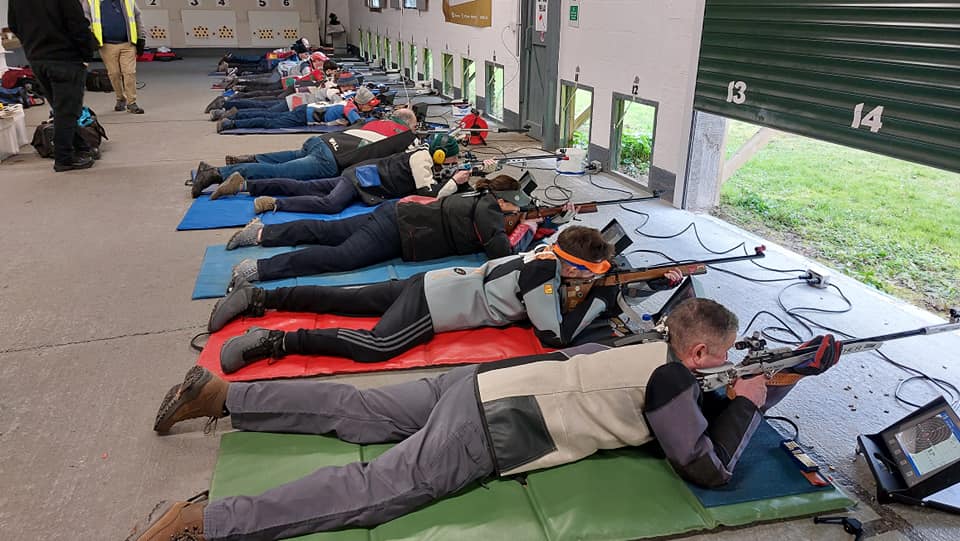 A group of floor shooters lie prone on the floor of an indoor firing point, aiming out through square holes in the wall at targets on the outdoor range. They wear canvas and leather jackets for support, some in bright colours. Monitors show the score from the electronic target system.