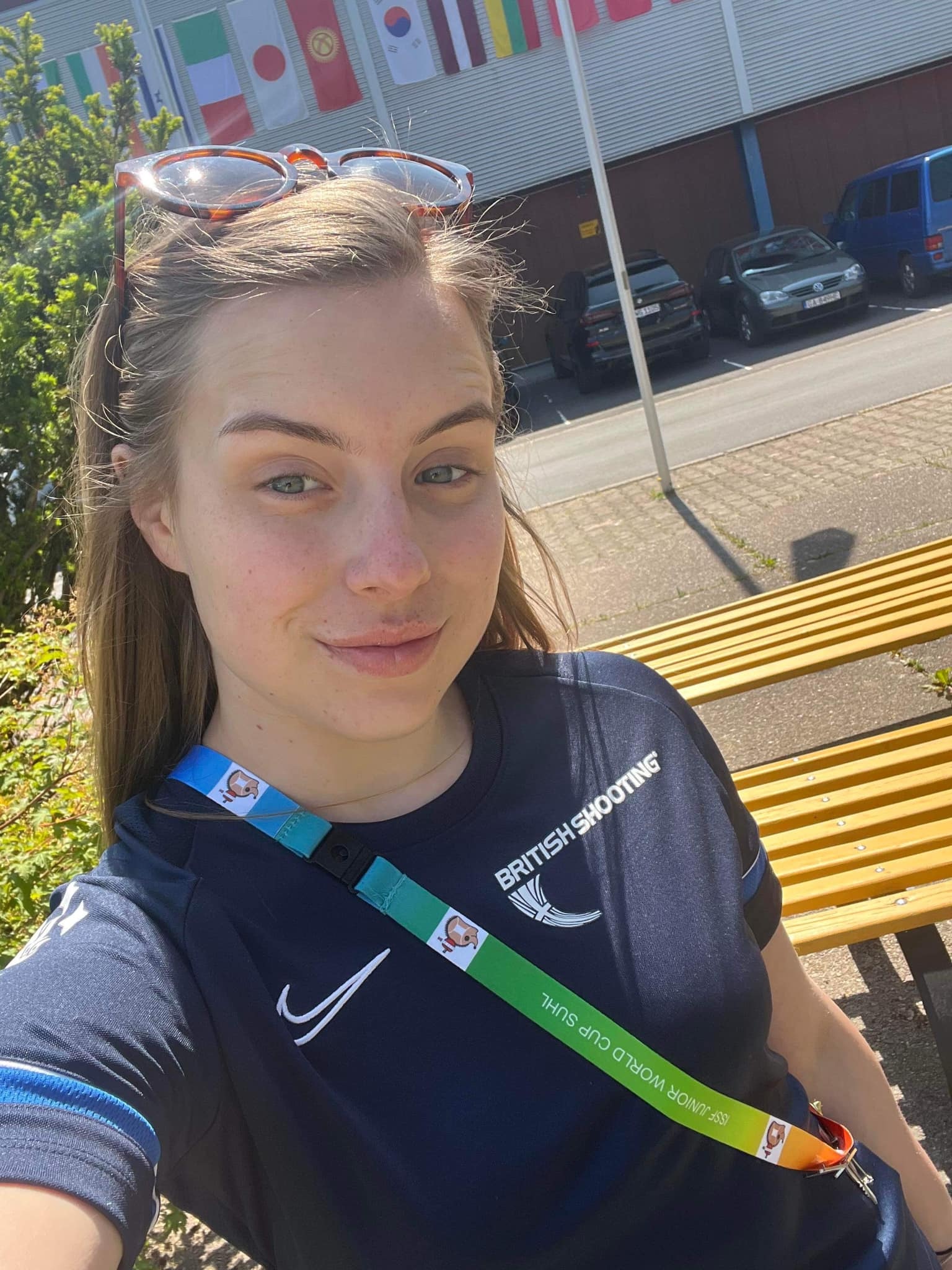 A young woman smiles at the camera as she takes a selfie. Her blue t-shirt features the British Shooting logo. A lanyard across her chest reads "ISSF Junior World Cup Suhl".