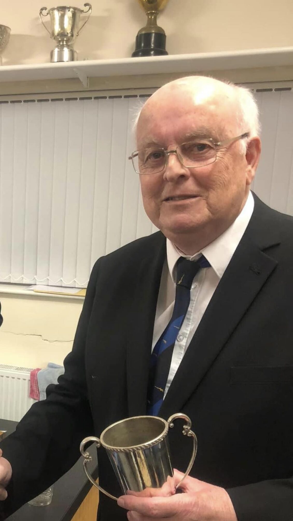 An older man in a black suit with a blue tie looks at the camera whilst holding a trophy.