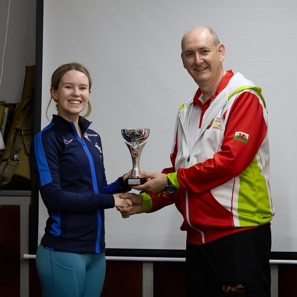 Two people stand in front of a projection screen. The man wearing a red, white and green top presents a prize to an athlete.