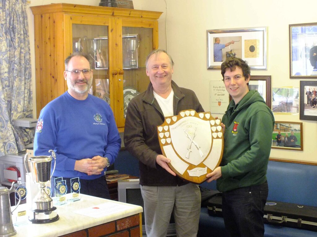 Three smiling people stand in the corner of a room. Two are holding a large wooden shield that they have received on behalf of a team. Other trophies are arrayed on a counter on the left.