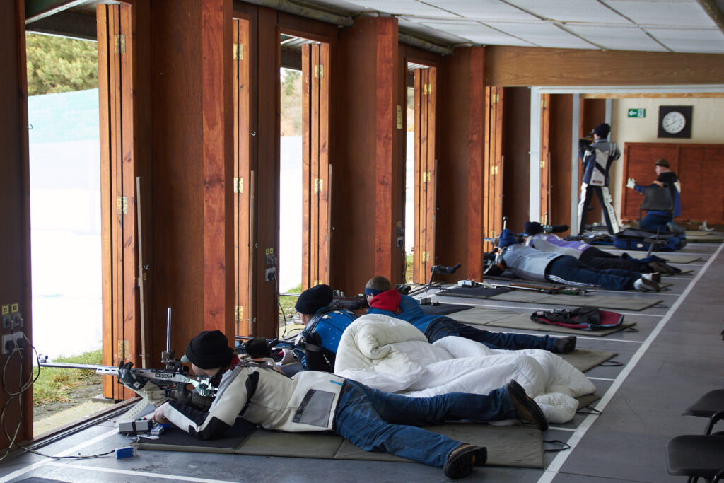 Target shooters on an indoor firing point. Most are lying down to shoot from the prone position. One is standing. The view outside is largely blown out to white as the snow reflects the sun brightly.