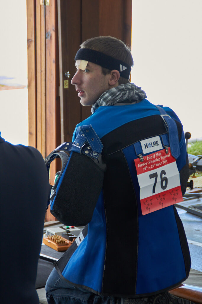 A young man kneels on the firing point. He wears a blue shooting jacket with a red and white sheet displaying his competitor number "76" attached to the back.