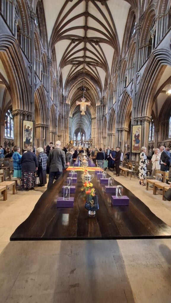 Heavy wooden tables are placed down the centre of a cathedral with a high vaulted ceiling. On the tables, various silver and crystal ware are arranged representing civic awards.