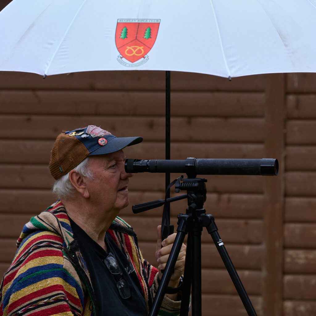 A man watches the shooting through a tripod-mounted spotting scope. He holds a white umbrella bearing the logo of Rugeley Rifle Club.