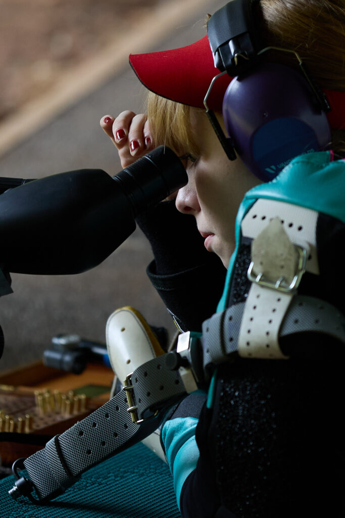 A young shooter adjusts their spotting scope before the start of a shooting match. They are wearing a red visor and blue ear defenders, along with a teal shooting jacket.
