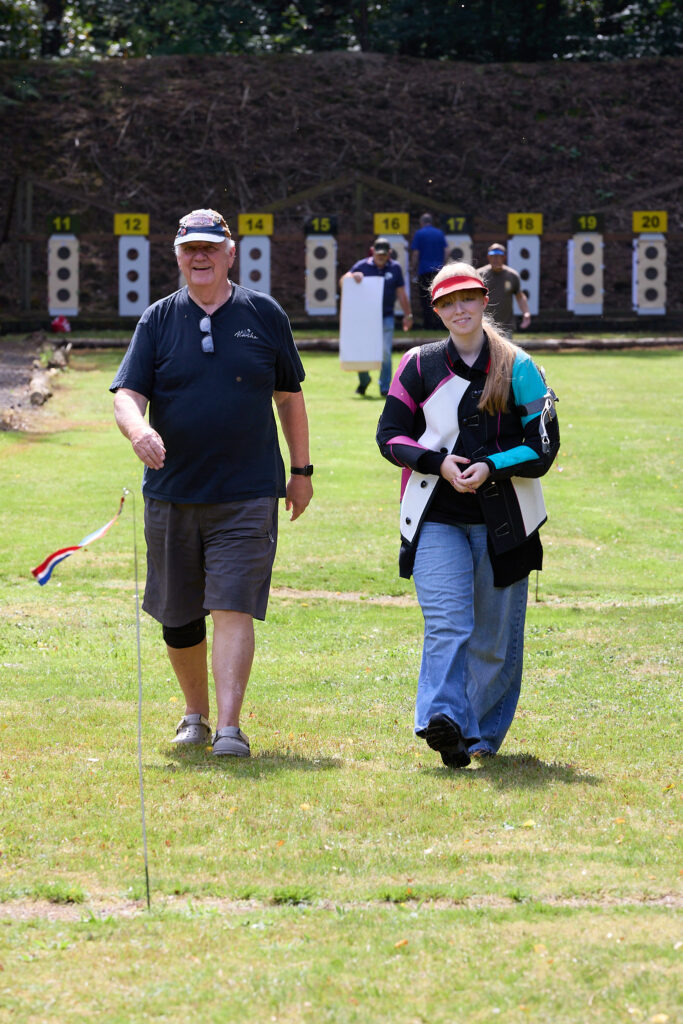 Two smiling shooters walk up a target shooting range after changing targets. One is wearing shorts and a blue t-shirt. The other is wearing jeans and a canvas shooting jacket in bright pink and cyan colours.