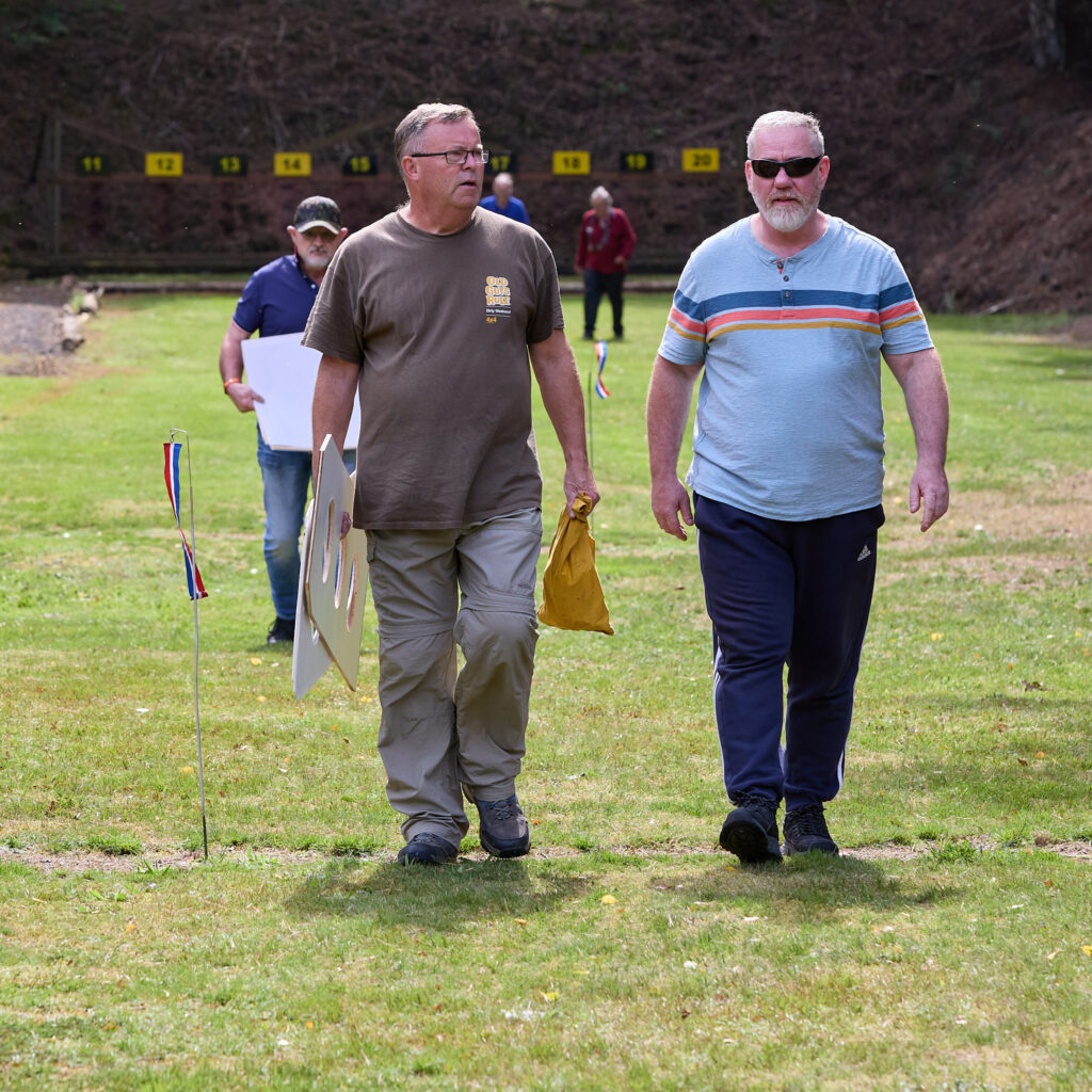 Two men walk up a shooting range, one carries wooden target frames and a yellow bag. A wind flag is visible to the left.