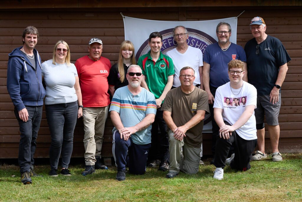 A group of target shooters stand and kneel in front of a shed, bearing a banner.