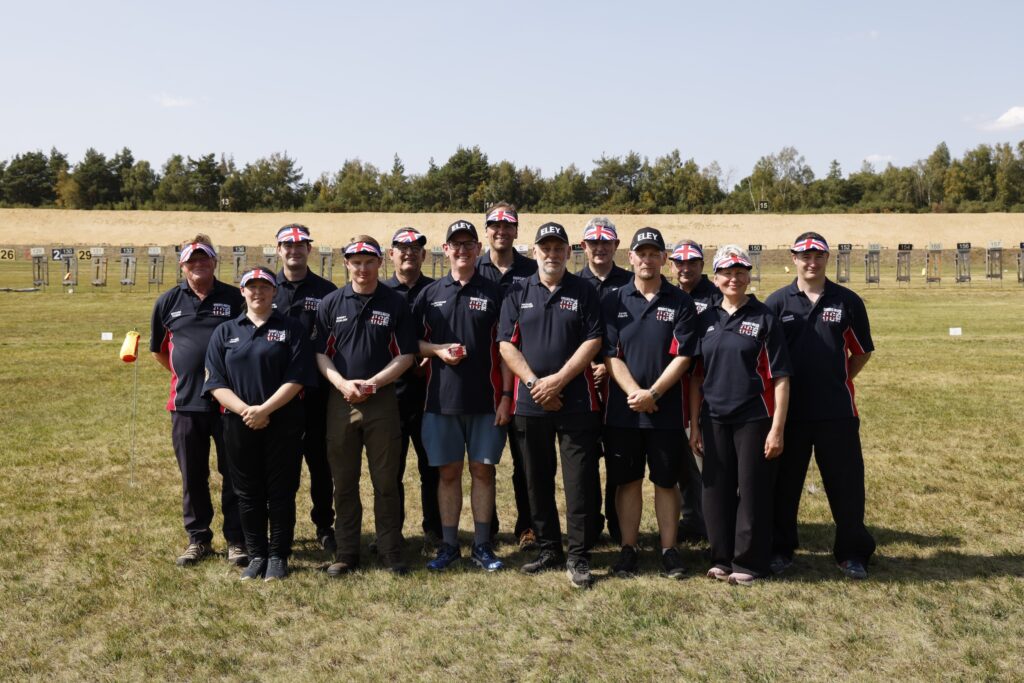 A group of smiling people pose as a group on a shooting range. They all wear visors bearing the union flag or black caps bearing the word "eley". Their shirts read