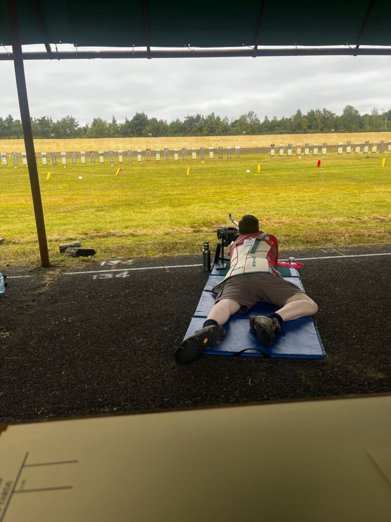 A man lies on a blue shooting mat aiming a rifle on a shooting range. A row of targets are visible in the distance. He is wearing brown shorts and a red and white shooting jacket.