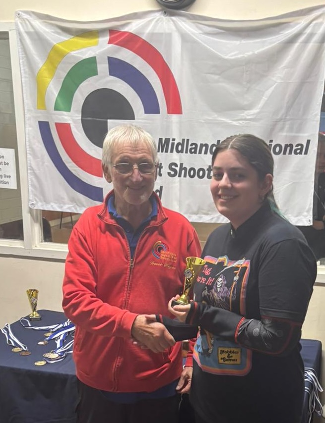 Two people stand in front of a table bearing trophies and medals. The man on the left is wearing a red jacket and presenting a trophy to the young woman on the right.