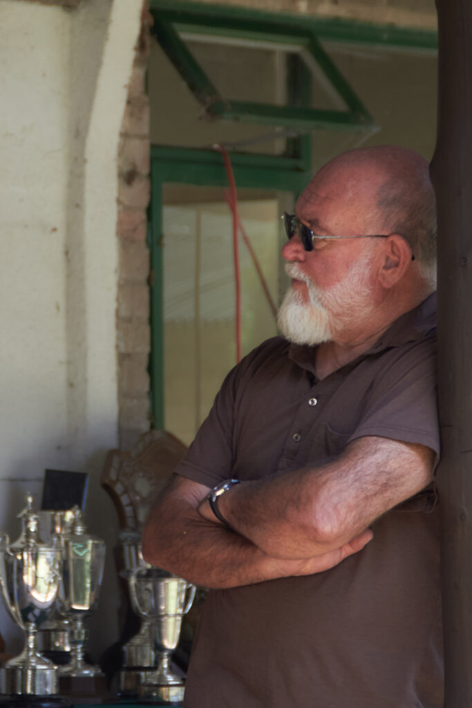 A man leans against a wooden post. He wears sunglasses and has a white beard. He wears a brown polo shirt. Behind him, a table is laden with silver trophies.